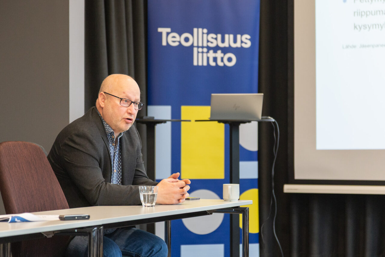 Henkilö istuu pöydän takana ja puhuu eteenpäin nojaten kädet pöydällä. Takana näkyy esitekangas, jossa lukee isoin kirjaimin Teollisuusliitto.