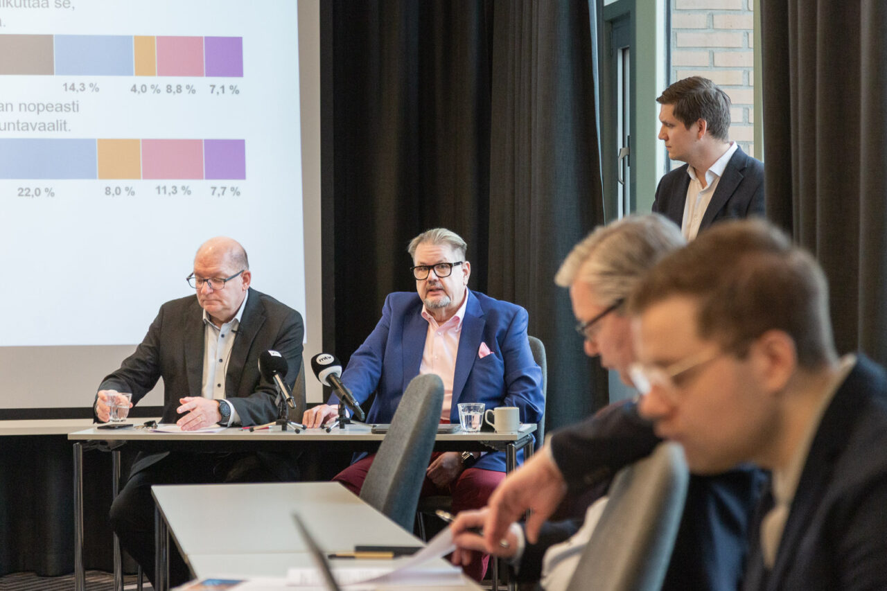Kaksi henkilöä istuu pöydän takana kameraa kohti. Toinen heistä puhuu ja toinen nostaa vesilasia. Kuvan etualalla on hankilöitä pöydän ääressä sivuttain puhujaan kohden.