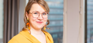 Läheltä kuvattu henkilö katsoo kameraan ja hymyilee. Takana näkyy ikkuna, josta näkyy kerrostalon seinä.