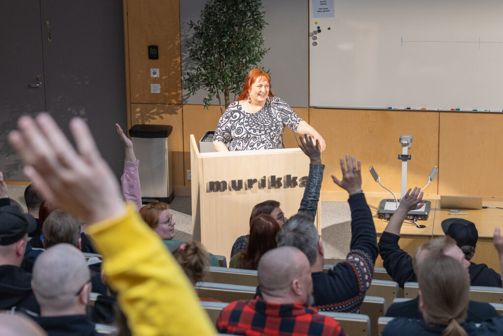 Hymyilevä henkilö seisoo puhujanpöntön takana luentosalin edessä. Salin takaa kuvatussa kuvassa näkyy ylös nostettuja käsiä.