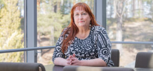 Henkilö istuu pöydän takana ja katsoo kameraan. Takana ikkunan läpi näkyy metsämaisema.