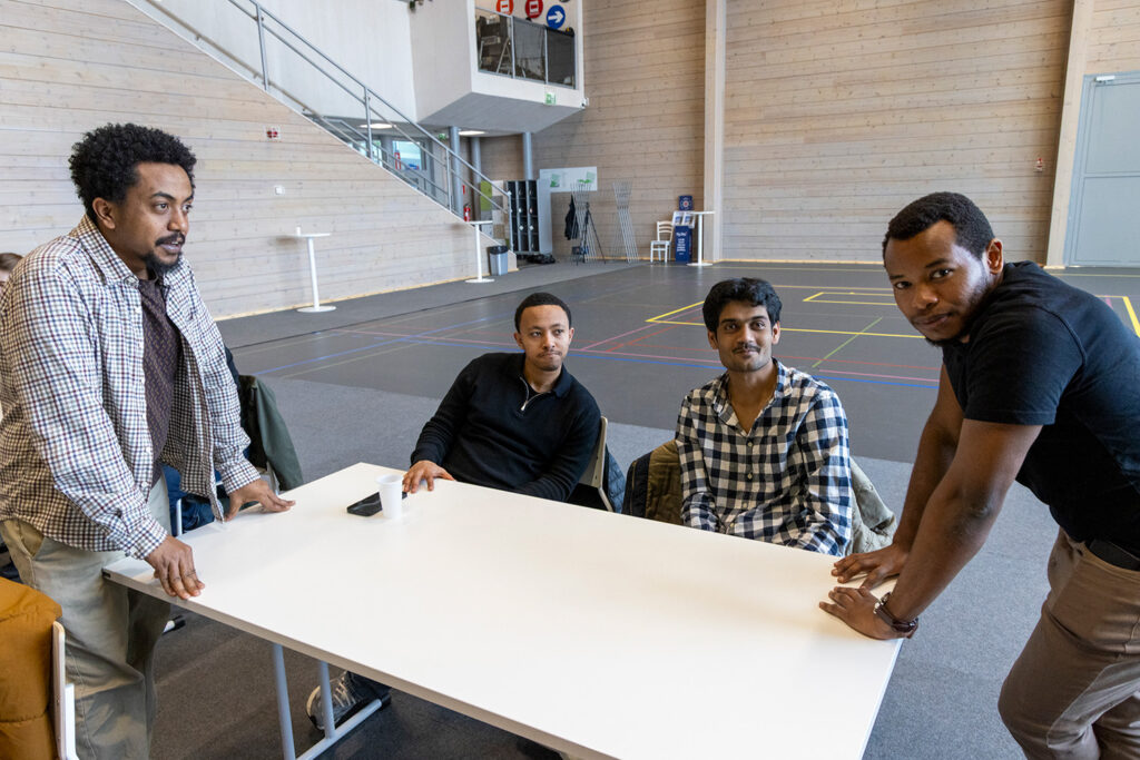 Neljä miestä on pöydän ympärillä. Etualalla kaksi miehistä seisoo ja kaksi istuu pöydän takana