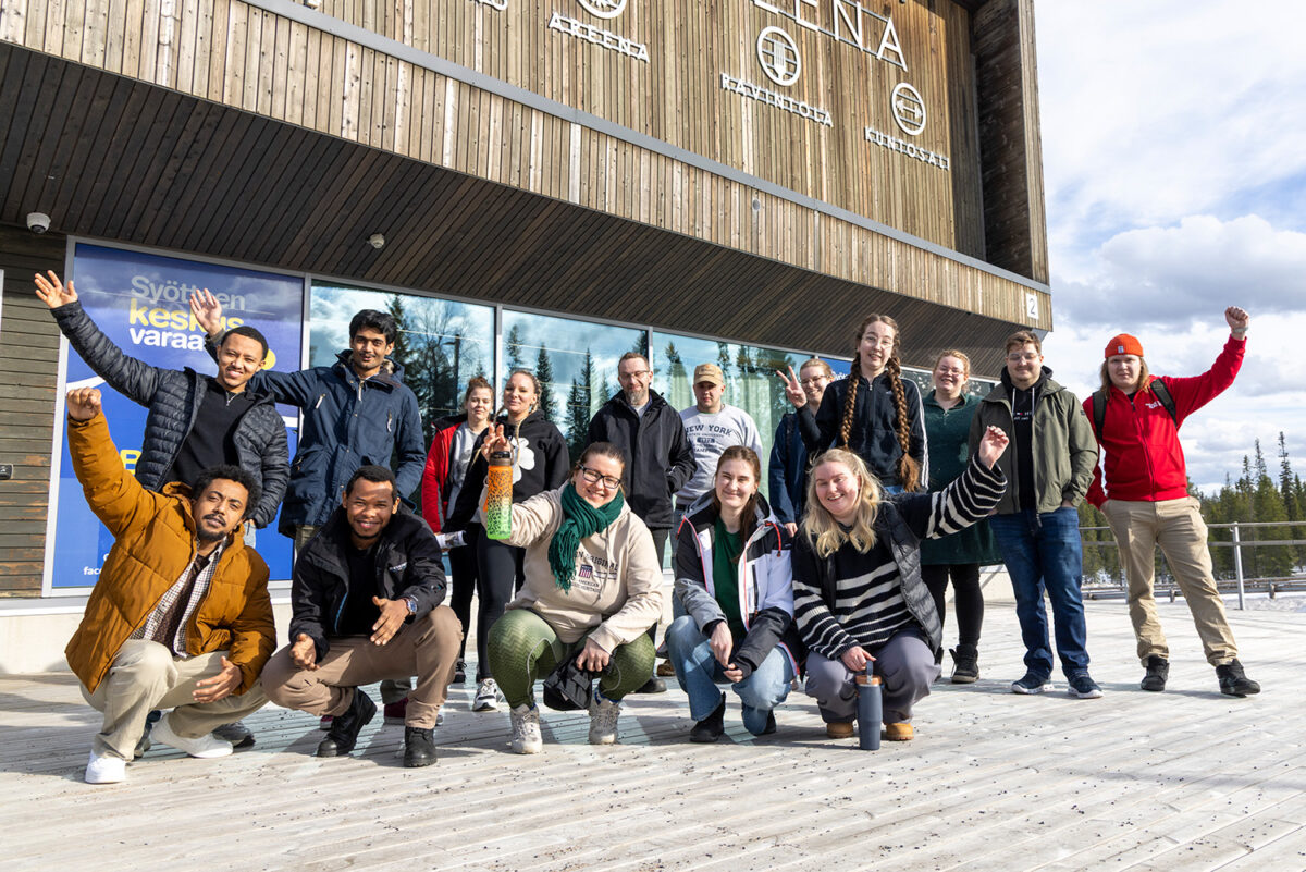 Ihmisiä ulkona rakennuksen edessä. Takana ihmiset seisovat ja etualalla ovat kyykyssä.