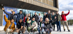 Ihmisiä ulkona rakennuksen edessä. Takana ihmiset seisovat ja etualalla ovat kyykyssä.