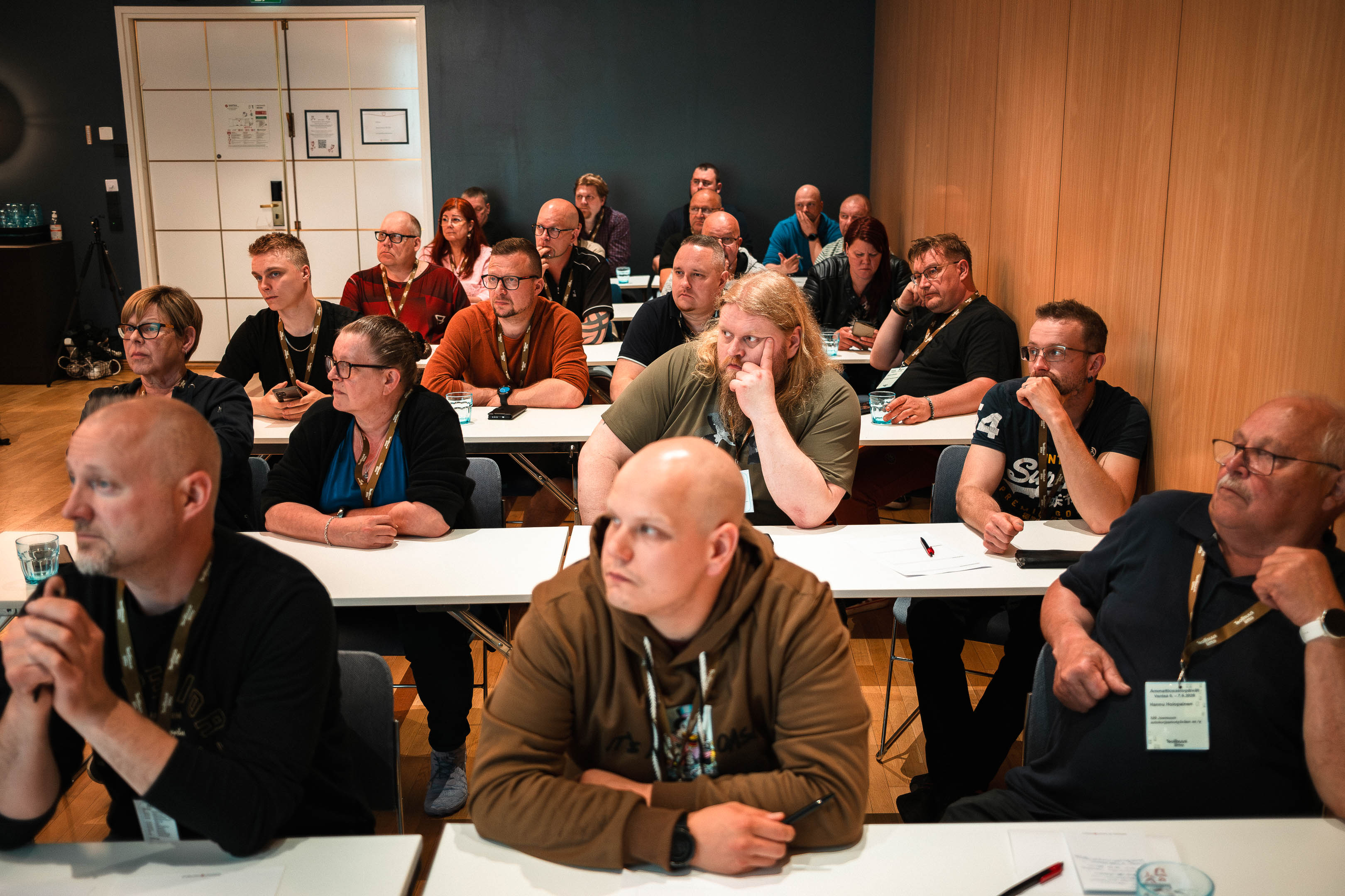 Ryhmä ihmisiä istuu riveissä valkoisten pöytien ääressä kokoustilassa ja katsoo eteenpäin. Osalla heistä on kaulassaan nimikortit.