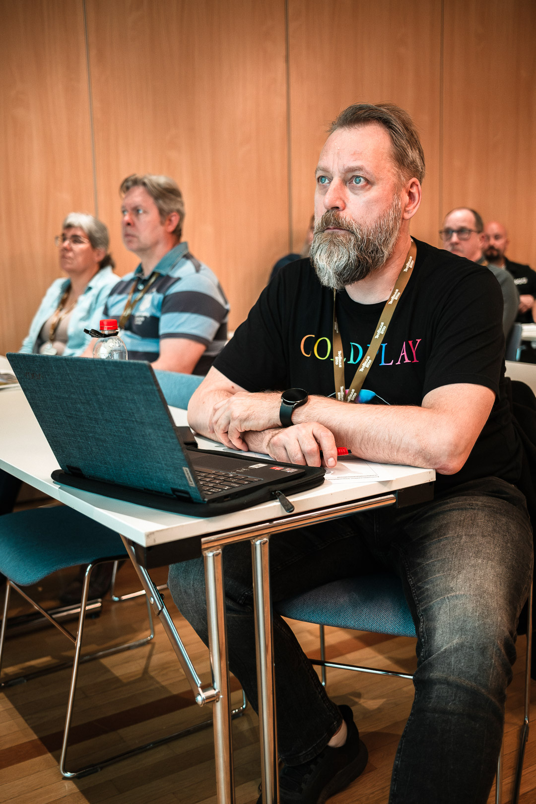 Parrakas mies istuu pöydän ääressä seminaarisalissa ja katsoo eteenpäin. Hänellä on yllään musta Coldplay-t-paita ja edessään kannettava tietokone. Taustalla näkyy muita yleisön jäseniä.
