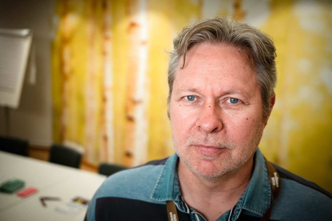 Lähikuva miehestä, jolla on siniset silmät ja harmahtavat hiukset. Hän katsoo suoraan kameraan ilmeettömänä. Taustalla on sumennettu keltainen seinä ja kokoushuone.