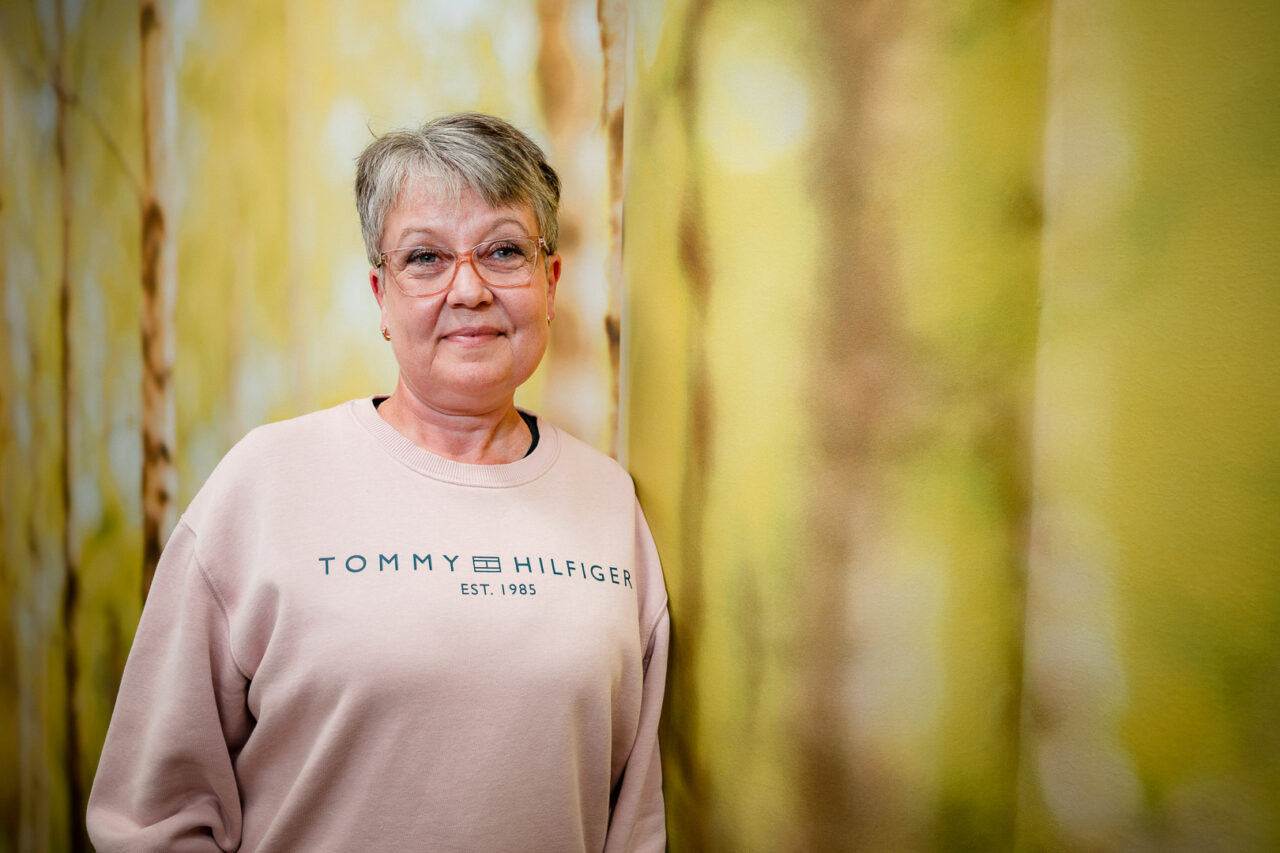 Hymyilevä, lyhyttukkainen nainen silmälaseissa ja vaaleanpunaisessa collegepaidassa nojaa kelta-vihreään, koivukuvioiseen seinään.