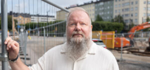 Henkilö pitää kättä työmaa-aitaa vasten. Takana näkyy kerrostaloja ja kaivinkone.