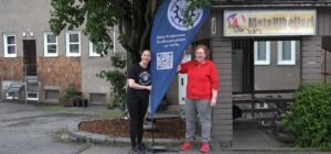 Kaksi henkilöä seisoo jalkakäytävällä. Heidän välissään on omalla jalalla seisova lippu, jossa lukee Etelä-Pirkanmaan Teollisuustyöväen ao 169 Ry.