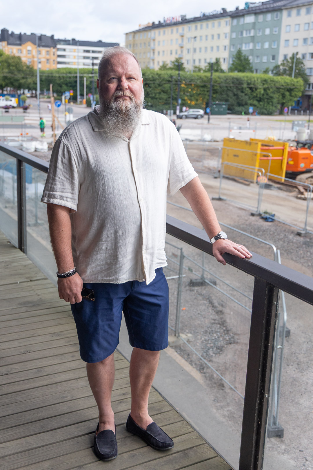 Henkilö seisoo katutason yläpuolella olevalla tasanteella ja nojaa kaiteeseen. Takana näkyy kaupunkimaisema, vihreitä puita ja maanrakennustyömaa. Henkilöllä on sortsit jalassaan.