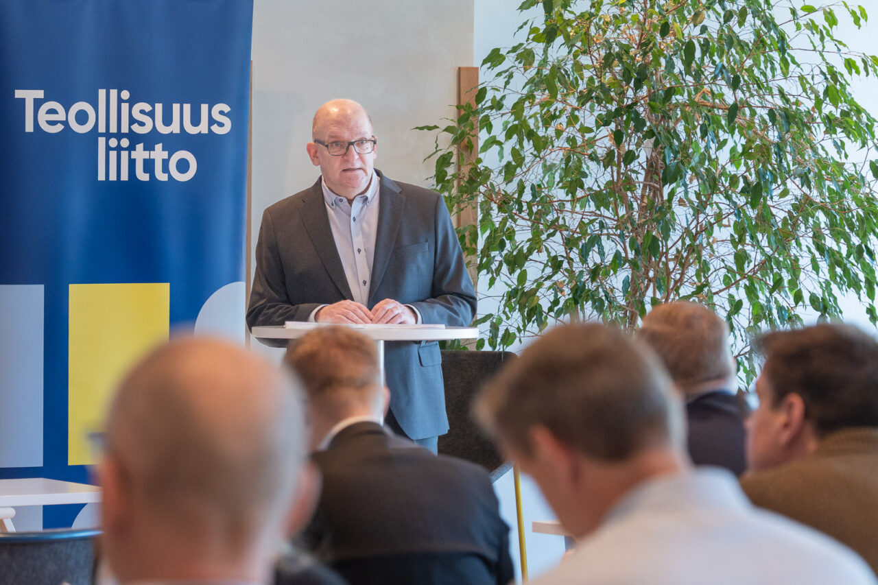 Henkilö puhuu pystypöydän takaa. Etualalla näkyy kuuntelijoiden hahmoja.