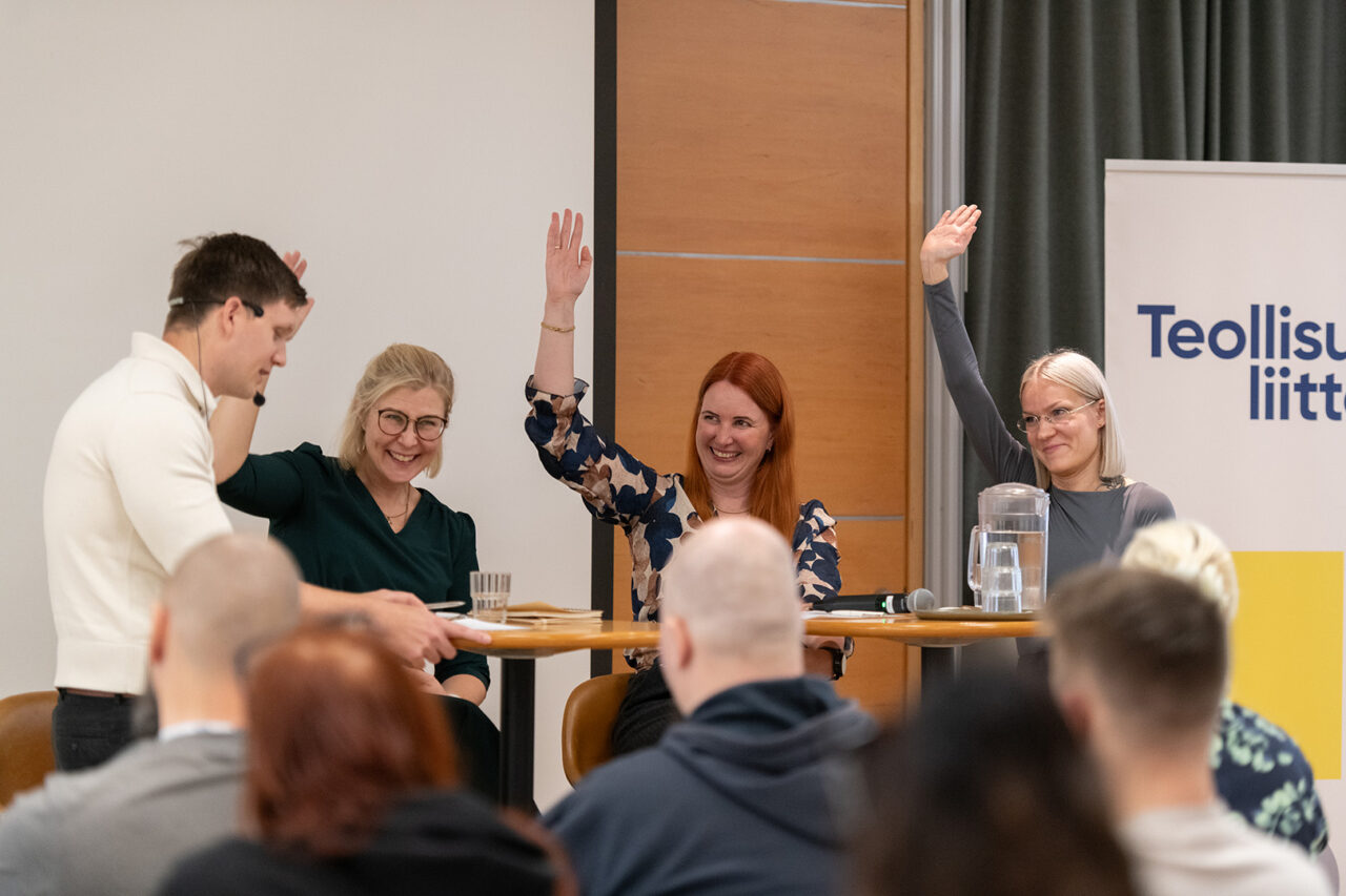 Kolme naista istuu ja hymyilee, mies seisoo pöydän ääressä.